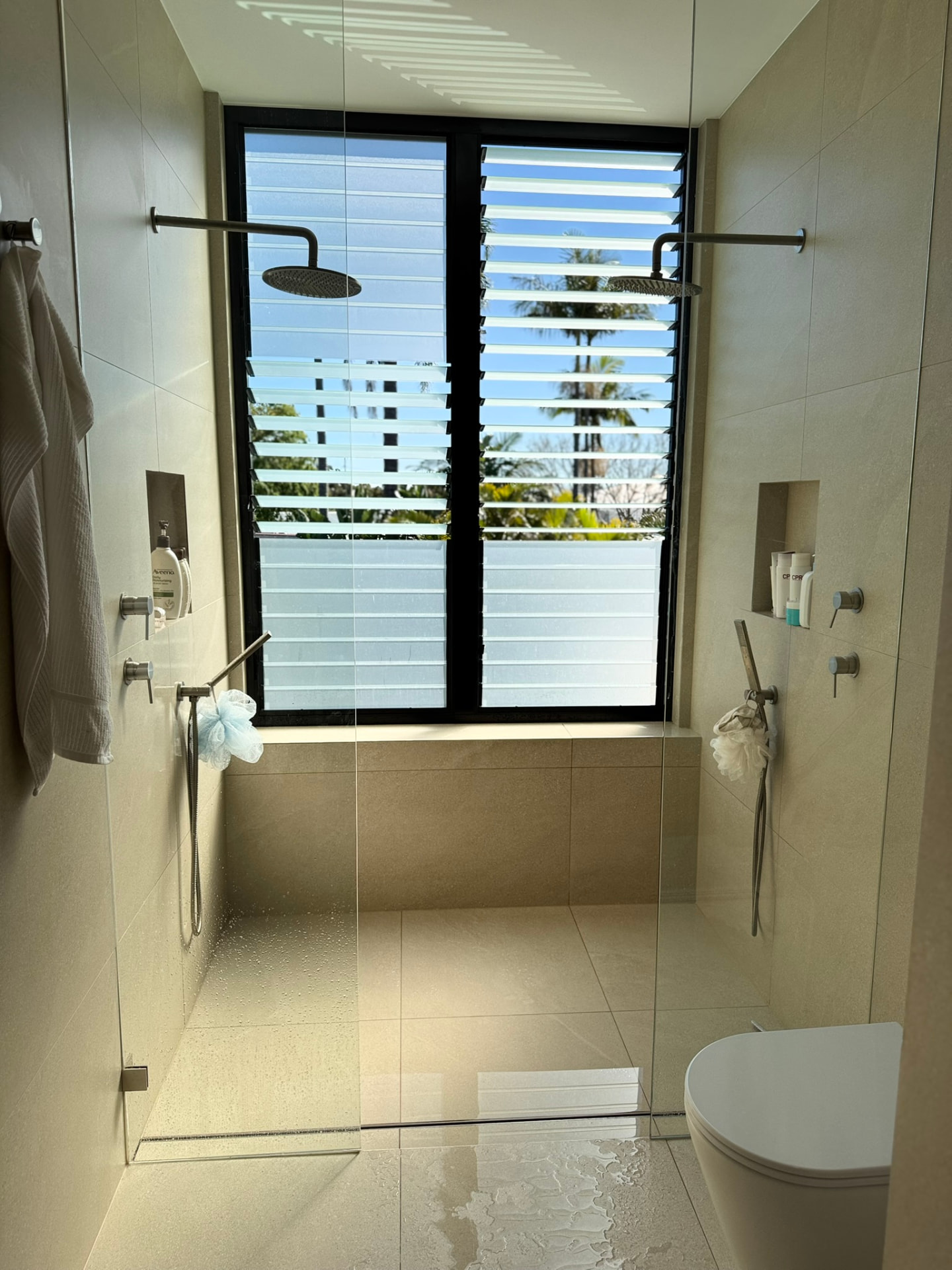 Twin rain-head shower in a wet area with a strip drain running across the entry under a louvre window