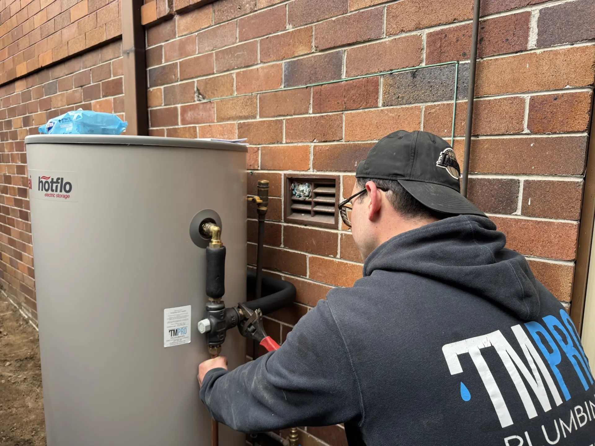 A new Rinnai Hotflo electric storage unit against a brick wall, with a plumber connecting the valves