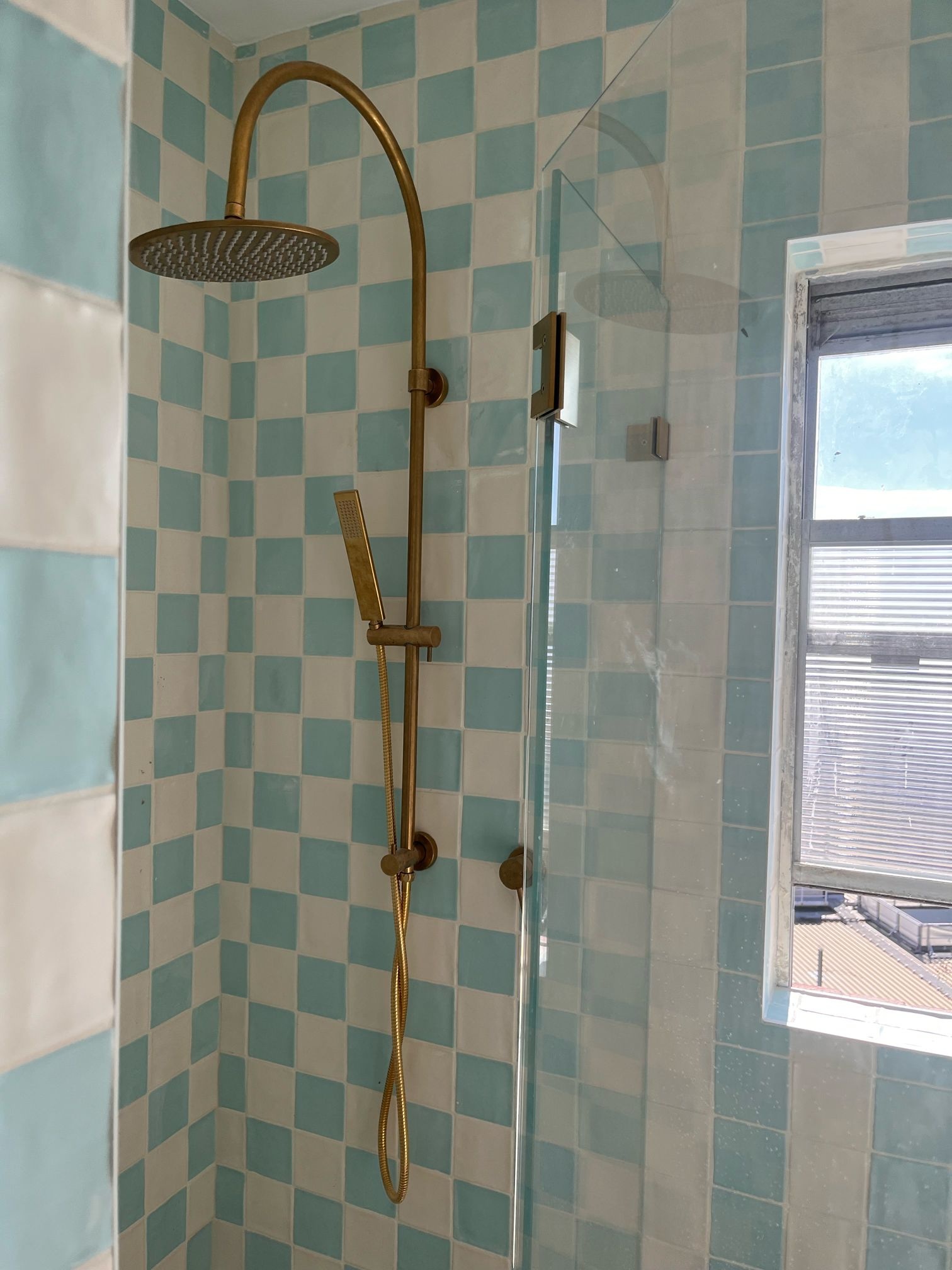 Brass showerhead and hand shower mounted over mint and white tiles beside a glass shower screen